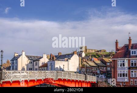 A part of Whitby town rising above a red painted bridge.  A public house in on the right and a church stands on the hill above. Stock Photo