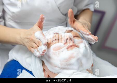 Close-up beautician doctor hands making anti-age procedures, applying foam cleansing mask for mid-aged female client at beauty clinic. Cosmetologist Stock Photo