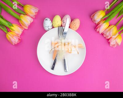 festive easter table cutlery with knife, fork, decorated with feather, bow, multi-colored eggs, yellow tulips flowers on pink, spring holiday background. Top view Stock Photo