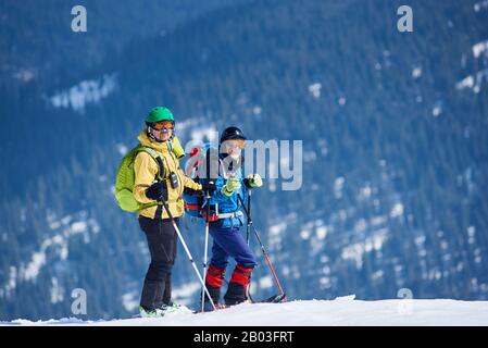 Two male skiers hikers in skiing equipment with backpacks on skis on background of mountain snowy slope covered with green spruce trees on frosty day. Winter tourism, ecological holidays concept. Stock Photo