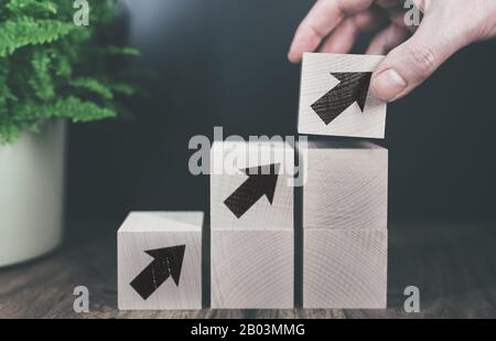 close-up of hand stacking wooden blocks with arrow symbol in steps, business or economic growth concept. Stock Photo
