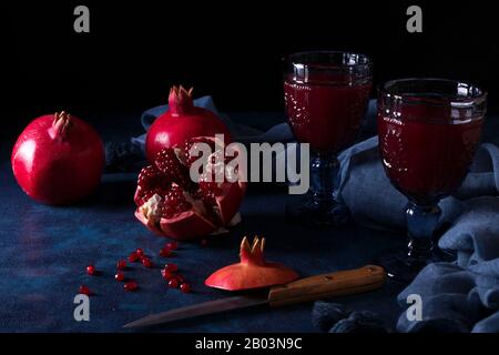 2 glasses of fresh pomegranate juice with few ripe real pomegranates on dark blue background. Partially peeled pomegranate, rouge seeds and peeling kn Stock Photo
