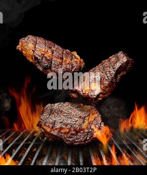 Delicious grilled pork or beef steaks are falling down on black background. Barbecue bbq grill, flaming fire, ember charcoal and smoke. Cooking and re Stock Photo