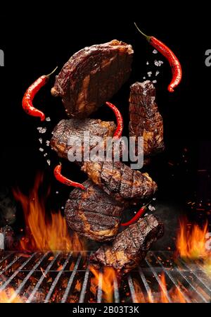 Grilled pork or beef steaks with chilli and salt are falling down on black background. Barbecue bbq grill, flaming fire, ember charcoal and smoke. Coo Stock Photo