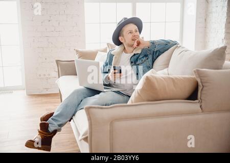 Bearded man coworker hipster in hat working living room home. Male sitting at sofa using laptop and mobile phone. Blurred light background Stock Photo