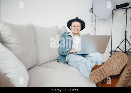 Freelancer photographer in hat video blogger works laptop, mounts videos for vlog about travel, in background lighting equipment Stock Photo
