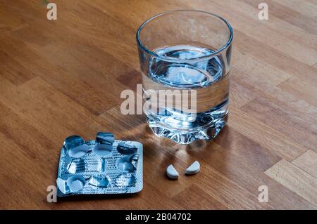 Painkiller tablet broken in half to administer half dosage amount, on bedside table with a glass of water. Stock Photo