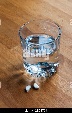 Painkiller tablet broken in half to administer half dosage amount, on bedside table with a glass of water. Stock Photo