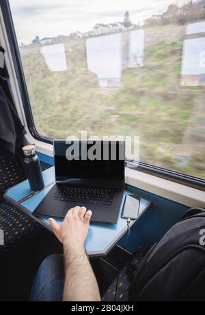 Man using laptop and charging smartphone inside train. Businessman commuting. Swiss high-speed train interior. Modern technology and public transport Stock Photo