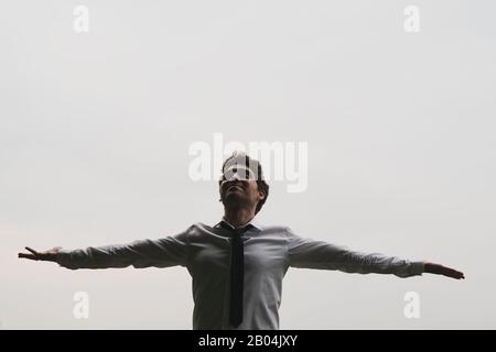 Young relaxed businessman celebrating success with his arms spread widely standing under grey sky. Stock Photo