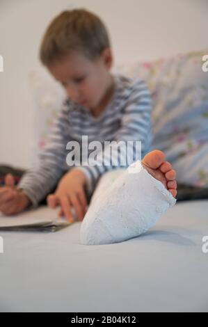 Low angle view of a toddler boy with broken leg in a cast, sitting on a bed drawing. Stock Photo