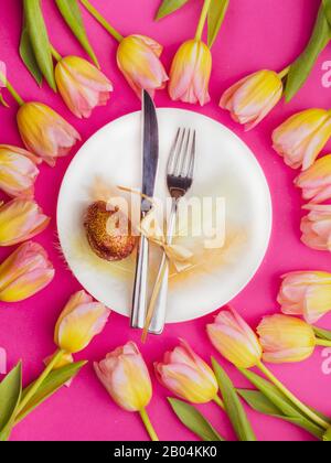 Beautiful luxury easter table cutlery settings as knife, fork with colorful tulips frame and gold painted eggs with glitters on pink surface. Holiday background, greeting card, close up Stock Photo