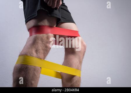 closeup, bottom view of the guys muscular legs in sports shorts, stretching fitness elastic bands with his legs, involvement of leg muscle groups that Stock Photo