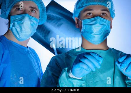 Portrait of two surgeons in sterile gowns, hats and masks. One surgeon, preparing to operate, holds a scalpel with a needle holder in his hands, anoth Stock Photo