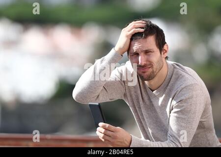 Confused man holding mobile phone complaining after mistake sitting in a rural town Stock Photo