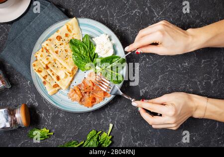 Female hands hold fork and knife. Crepes with smoked salmon, spinach and sour cream on stone background. Top view Stock Photo