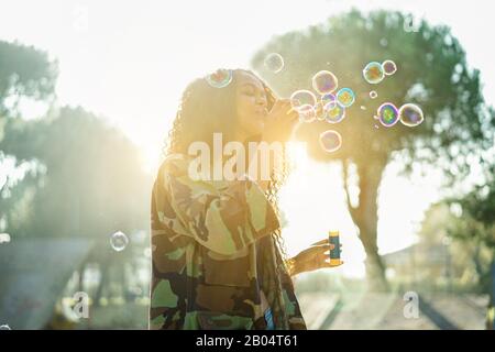 Young afro hair woman blowing soap bubble in city skate park with back sun light - Portrait of african happy girl having fun outdoor - Happiness conce Stock Photo