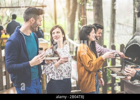 Group of happy friends cooking and eating at barbecue dinner outdoor - Young people toasting wine for bbq in wood house backyard - Friendship concept Stock Photo