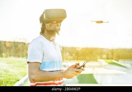 Young man using a drone with remote controller wearing virtual reality glasses making photos and videos - Young guy having fun with new technology tre Stock Photo