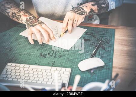 Tattoo artist cutting paper flower illustration inside ink studio - Hipster tattoer at work - New body skin trends generation - Contrast vintage filte Stock Photo