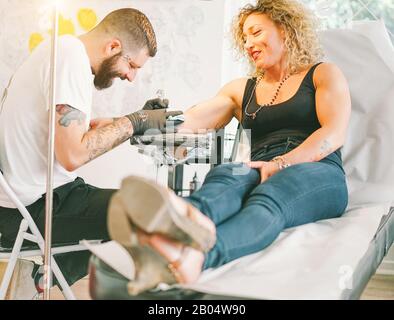 Bearded tattoo artist making tattoo inside ink studio on blond woman - Tattooer at work - Contemporary trend fashion concept - Main focus on female fa Stock Photo