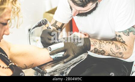 Bearded tattoo artist making tattoo inside ink studio on young blond woman - Tattooer at work - Skin trend fashion concept - Focus on man hand machine Stock Photo