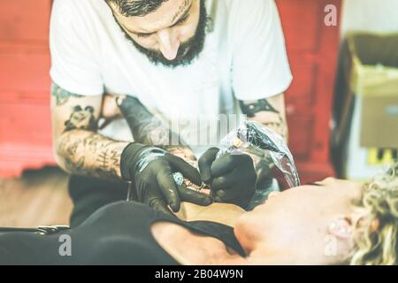Bearded tattoo artist making tattoo inside ink studio on young blond woman - Tattooer at work - Skin trend fashion concept - Focus on man hands machin Stock Photo