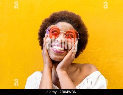 Fashion hippie african woman smiling and wearing sunglasses with yellow ochre wall in background - Black afro girl having fun - Focus on face - Youth Stock Photo