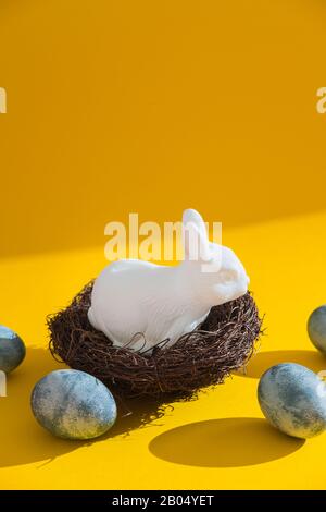Blue textured colored eggs and white plastic easter bunny sitting in a nest against a bright yellow background. Hard light Stock Photo