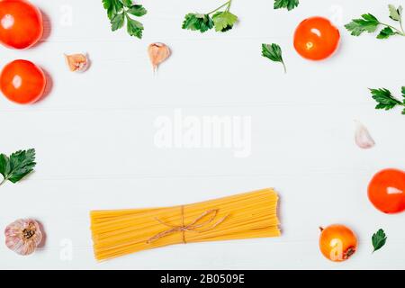 Flat lay frame of bunch uncooked dry spaghetti next to fresh parsley leaves, onion, garlic and tomatoes on white background. Stock Photo