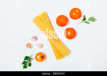 Flat lay food arrangement of spaghetti pasta next to fresh tomatoes, onions, garlic and parsley leaves on white table. Stock Photo
