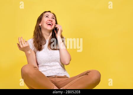 A girl in a T-shirt on a yellow background is talking on the phone with a friend and laughs out loud. The concept is nice communication. Close up. Stock Photo