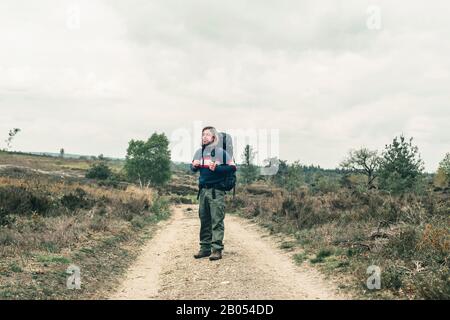 Man with long blonde hair and red backpack on dirt road in hilly landscape under cloudy sky. Stock Photo