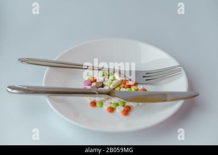 Pills on a white plate. The gesture with a knife and fork is simply excellent, on a plate with pills. A pile of multi-colored tablets disguised as Stock Photo