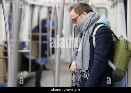 Tired man in eyeglasses in metro train. Young exhausted male standing and sleeping in subway after work, people on background. Fatigue, sleep deprivat Stock Photo