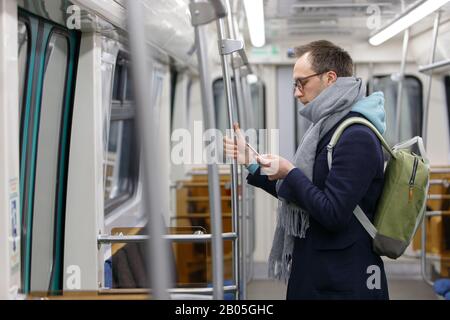 Caucasian young man passenger in eyeglasses with backpack holding handrail, using and looking at smart mobile phone at underground train. Subway Stock Photo