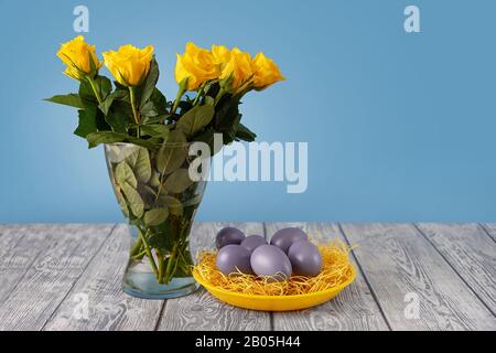 Yellow roses in a vase next to a yellow plate with blue Easter eggs Stock Photo