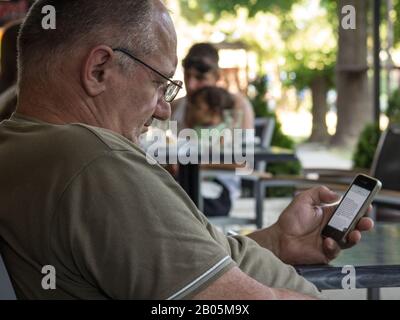 ALIBUNAR, SERBIA - JUNE 6, 2015: Oldd sernior white man sitting in a cafe looking at an Apple IPhone 4 smartphone using the mobile internet data.   Pi Stock Photo