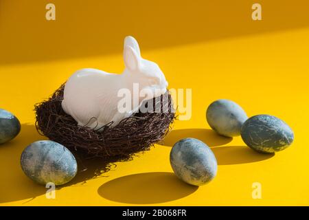 Blue textured colored eggs and white plastic easter bunny sitting in a nest against a bright yellow background. Hard light Stock Photo
