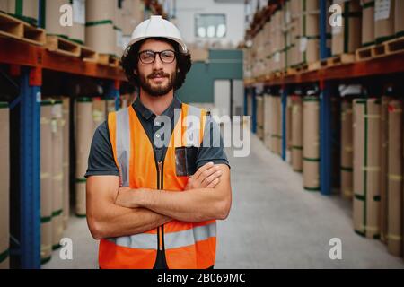 Portrait of confident worker wearing helmet and spectacles with orange vest uniform standing in aisle of heavy duty factory warehouse Stock Photo