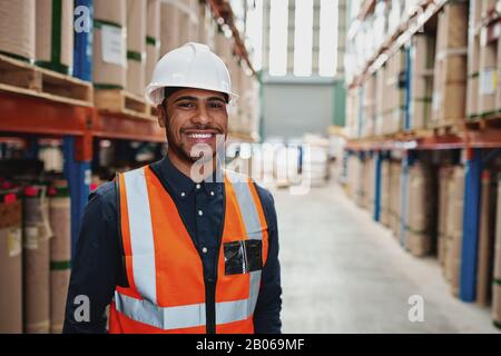 Happy male supervisor in warehouse standing in uniform with white hardhat smiling looking at camera - copy space on the right Stock Photo