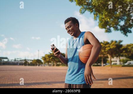 Happy young ,ale sportive basketball player holding ball in his arms using mobile phone at park Stock Photo