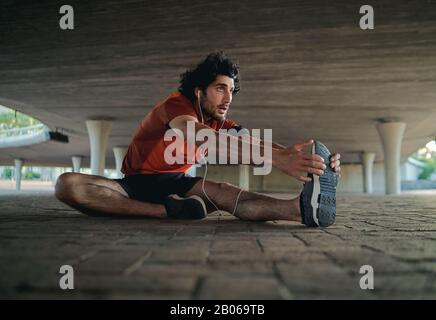 Fit young man in sportswear sitting on pavement under the bridge warming up and doing stretching exercise before going for a run Stock Photo
