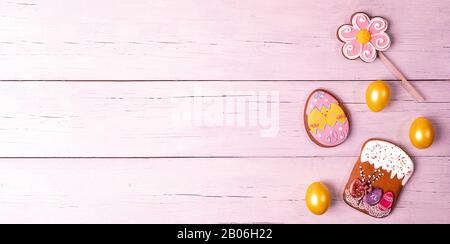 Festive Easter banner or greeting card. Painted yellow eggs, colorful gingerbreads in form of Easter cake, flower and funny egg on pink toned wooden b Stock Photo
