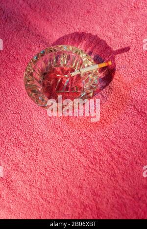 Ashtray with burning cigarette on sunbed with pink towel. Stock Photo