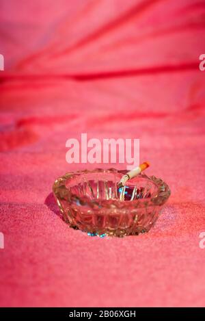 Ashtray with burning cigarette on sunbed with pink towel. Stock Photo