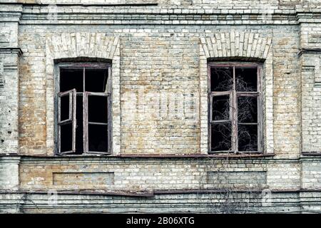 Old shabby ruined abandoned brick building wall facade with two wooden brocken glass windows and dark black background. Shattered grunge aged house Stock Photo
