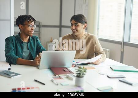 Two young businessmen in casual clothing sitting at the table talking and using laptop in work at office Stock Photo