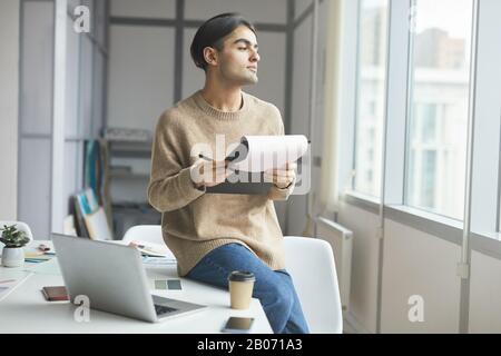 Pensive young businessman sitting on his workplace and looking at window while reading a business document at office Stock Photo
