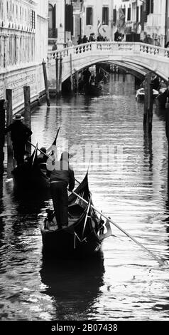 City of Venice, Italy, Europe.  Black & White image of Gondoliers rowing towards stone bridge along narrow canal Stock Photo